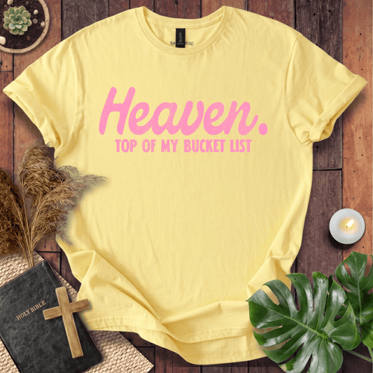 A pale yellow short-sleeve T-shirt laid flat on a rustic wooden surface, printed with the words 'Heaven. TOP OF MY BUCKET LIST' in pink.
