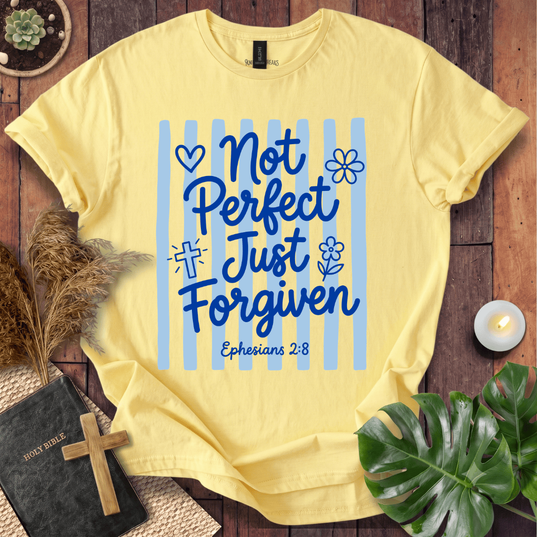 A pale yellow graphic T-shirt laid flat on a rustic wood surface with decorative props including a Bible, cross, pampas grass, candle, and green leaves. The shirt has blue vertical stripes and the text 'Not Perfect Just Just Forgiven Ephesians 2:8' with small heart, flower, and cross icons.