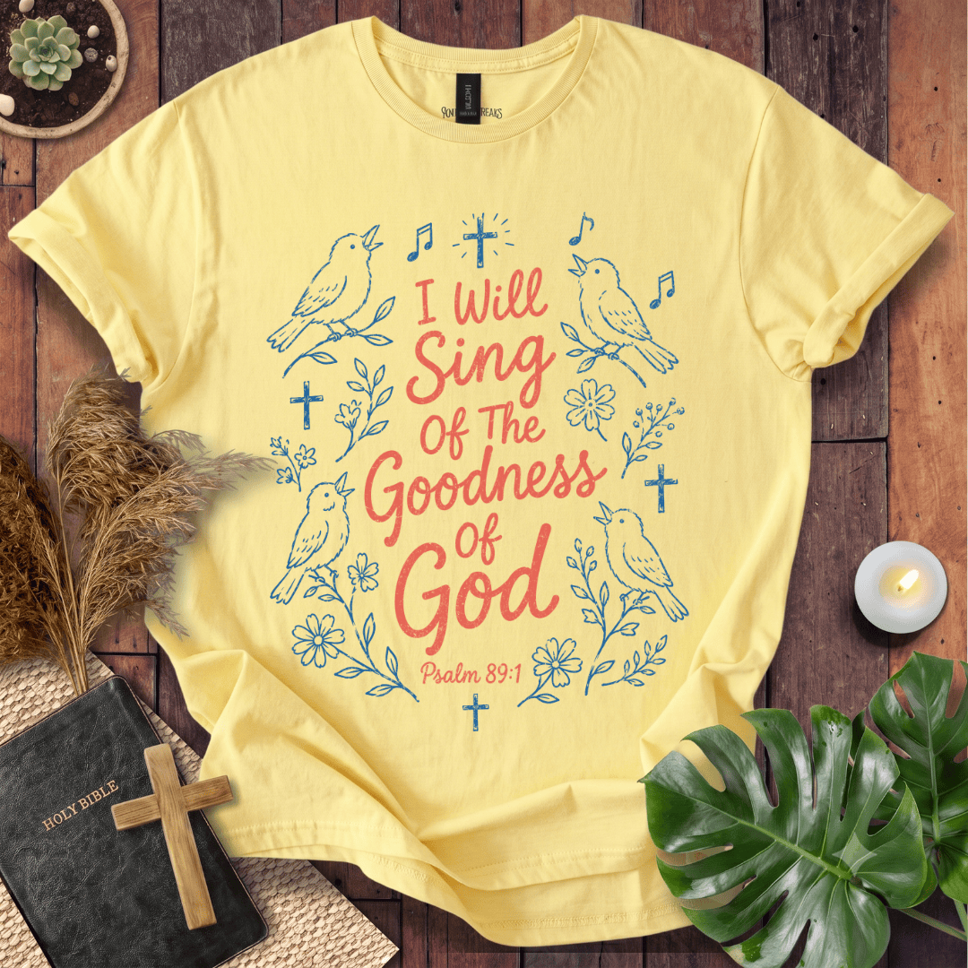 A pale yellow graphic T-shirt laid flat on a rustic wooden surface, surrounded by decorative props including a small plant, dried grass, a candle, a Bible with a wooden cross, and green leaves. The shirt features blue line drawings of birds, flowers, and crosses with coral-red text reading, 'I Will Sing Of The Goodness of God Psalm 89:1'.