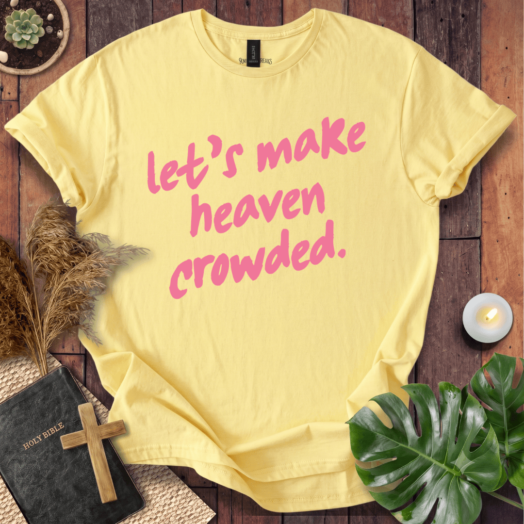 A pale yellow short-sleeve T-shirt laid flat on a rustic wooden surface, printed with the phrase "let’s make heaven crowded." in pink script.