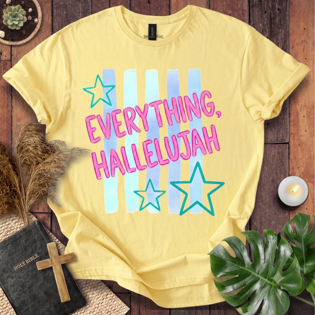 A pale yellow graphic T-shirt laid flat on a rustic wood surface, surrounded by decorative props including a small plant, dried grass, a candle, a Bible with a cross, and a green leaf. The shirt has pastel vertical stripes and the text 'EVERYTHING, HALLELUJAH' with outlined stars.