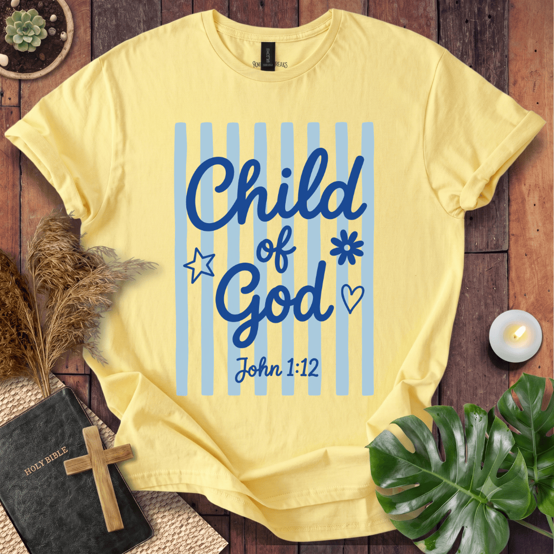 A pale yellow graphic T-shirt laid flat on a rustic wood surface, surrounded by decorative props including a small book with a cross, dried grass, a candle, and green leaves. The shirt has blue vertical stripes and the text 'Child of God John 1:12' with simple blue icons.
