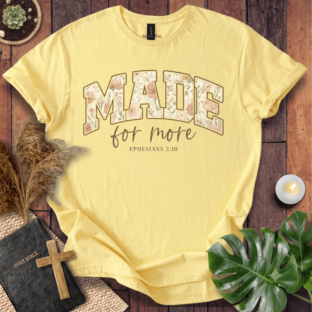 A pale yellow graphic T-shirt laid flat on a rustic wooden surface with decorative props including a Bible, cross, dried grass, candle, plant leaves, and a small succulent. The shirt has large floral-filled lettering reading “MADE” with smaller script text “for more” and “EPHESIANS 2:10” underneath.