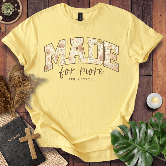 A pale yellow graphic T-shirt laid flat on a rustic wooden surface with decorative props including a Bible, cross, dried grass, candle, plant leaves, and a small succulent. The shirt has large floral-filled lettering reading “MADE” with smaller script text “for more” and “EPHESIANS 2:10” underneath.