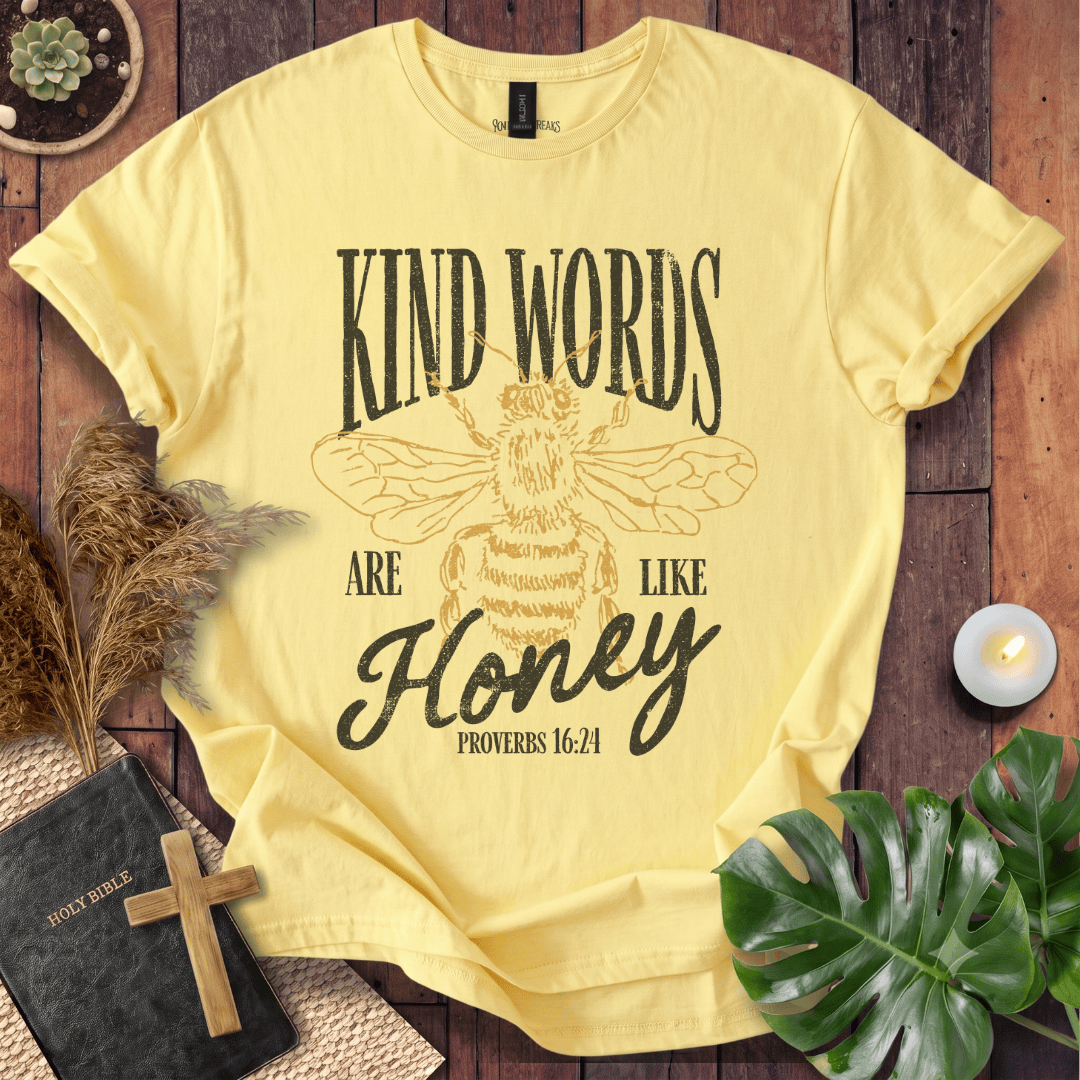 A pale yellow graphic T-shirt laid flat on a rustic wood surface, surrounded by decorative props including a Bible with a cross, dried grass, a small candle, and green leaves. The shirt has a large bee illustration and the text 'KIND WORDS ARE LIKE Honey PROVERBS 16:24' printed on the front.
