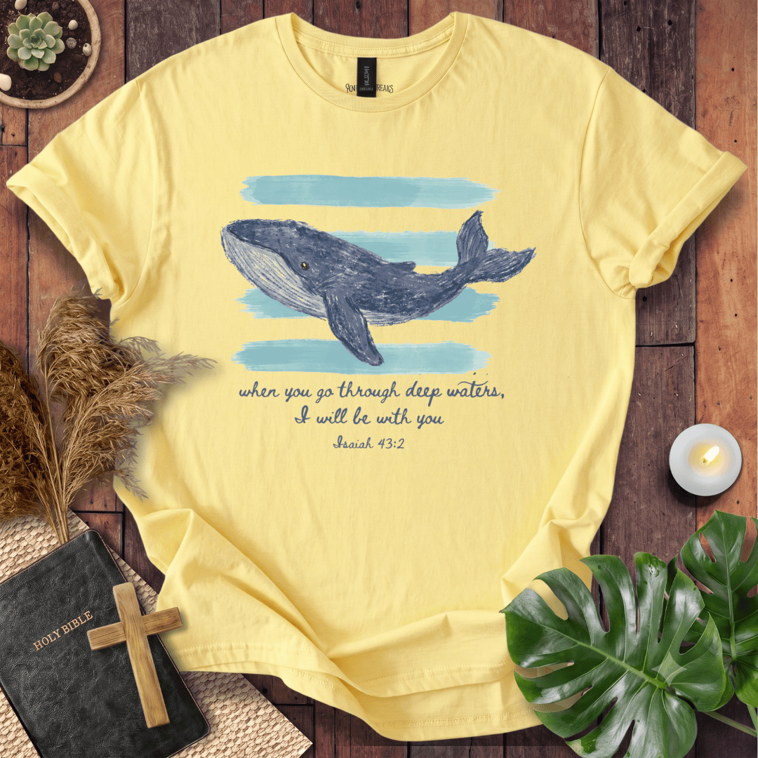 A pale yellow graphic T-shirt laid flat on a rustic wood surface, featuring a blue whale illustration, light blue watercolor bands, and the printed phrase 'when you go through deep waters, I will be with you' with 'Isaiah 43:2' below. Decorative props including a Bible, cross, dried grass, candle, and green leaves surround the shirt.