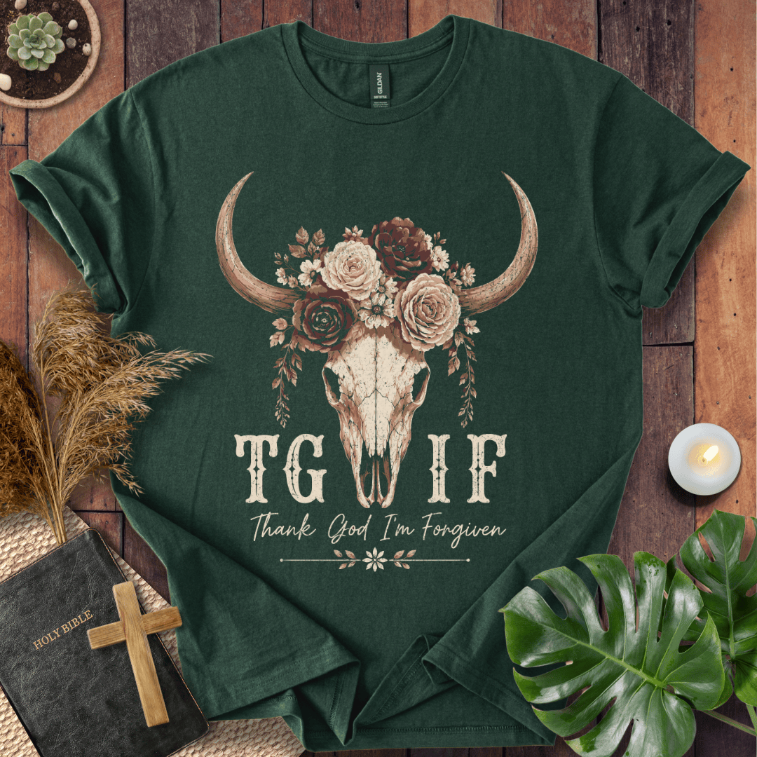 A dark forest green graphic t-shirt laid flat on a rustic wooden background, surrounded by decorative props including a bible, a wooden cross, a lit candle, dried grass, and a monstera leaf. The t-shirt features a central graphic of a bull skull decorated with a floral wreath, with the text 'TG IF' and 'Thank God I'm Forgiven' printed below.