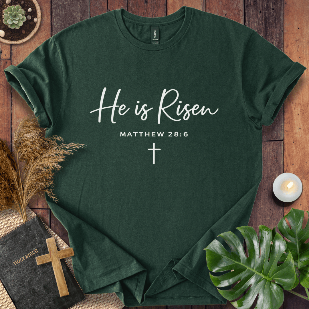 Easter Christian t-shirt with white "He is Risen" script and cross, styled on wood with a Bible and wooden cross.