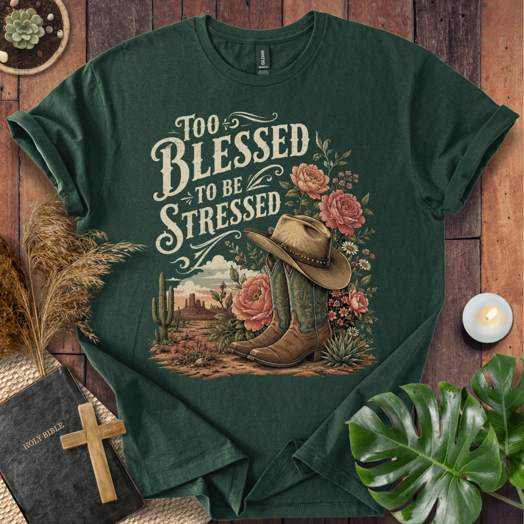 A dark green graphic t-shirt laid flat on a rustic wooden surface, surrounded by decorative props including a candle, dried grass, a monstera leaf, a bible with a wooden cross, and succulents. The t-shirt features a western-themed graphic with the text 'Too Blessed to be Stressed' and an illustration of a cowboy hat, boots, flowers, and a desert landscape.
