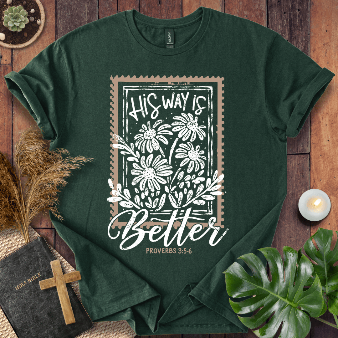 A dark green graphic t-shirt laid flat on a rustic wooden background, surrounded by decorative props including a Bible, a wooden cross, dried grass, a small plant, and a lit candle. The t-shirt features a white floral stamp-style graphic with the text 'HIS WAY IS Better PROVERBS 3:5-6'.