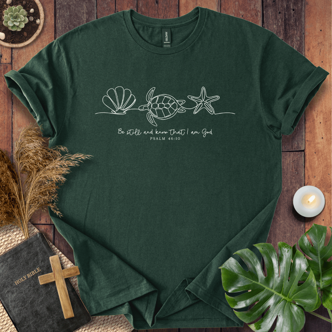 A dark green crewneck t-shirt with a white line art graphic of a seashell, sea turtle, and starfish, with the text 'Be still and know that I am God PHILIPPIANS 4:4' printed on the chest. The shirt is displayed in a flat-lay arrangement on a wooden surface with a Bible, a wooden cross, dried grass, a small candle, and a green monstera leaf.