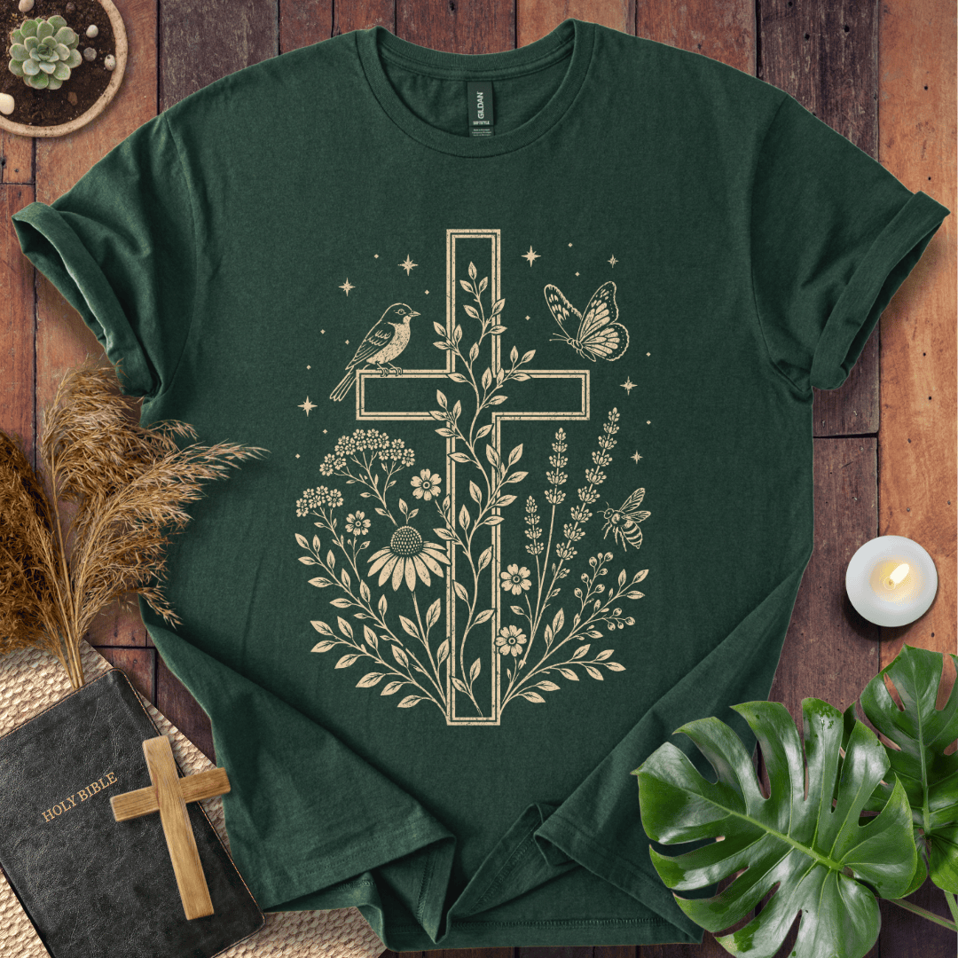 A dark green short-sleeve T-shirt laid flat on a rustic wooden surface, featuring a large cream-colored cross graphic with flowers, leaves, birds, and butterflies. The shirt is styled with a Bible, a small wooden cross, pampas grass, a lit candle, and green leaves.