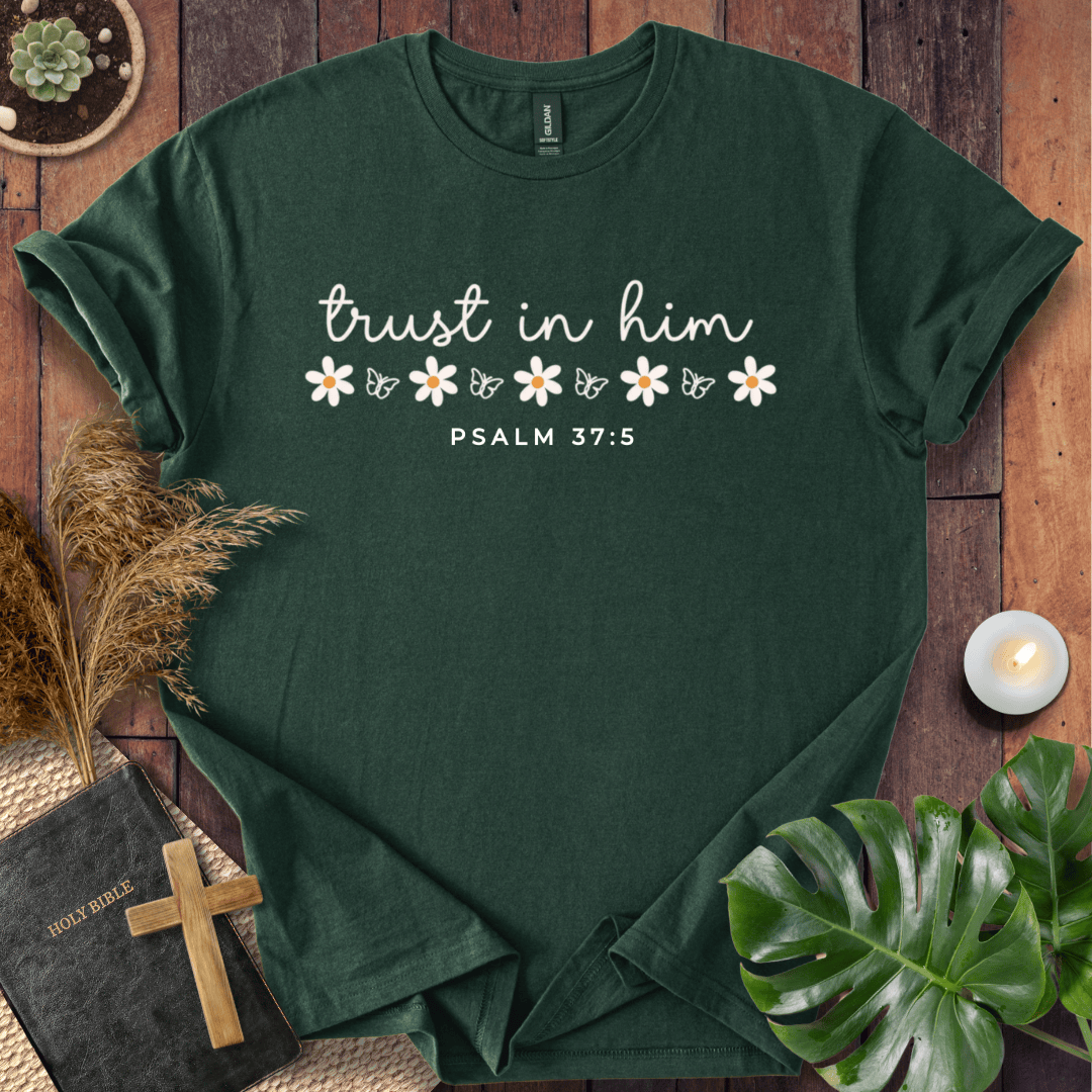 A dark green short-sleeve T-shirt laid flat on a rustic wooden surface, printed with the phrase 'trust in him' in white script, small white flowers, and 'PSALM 37:5' below. The shirt is styled with a book, cross, dried grass, a small candle, and green leaves around it.