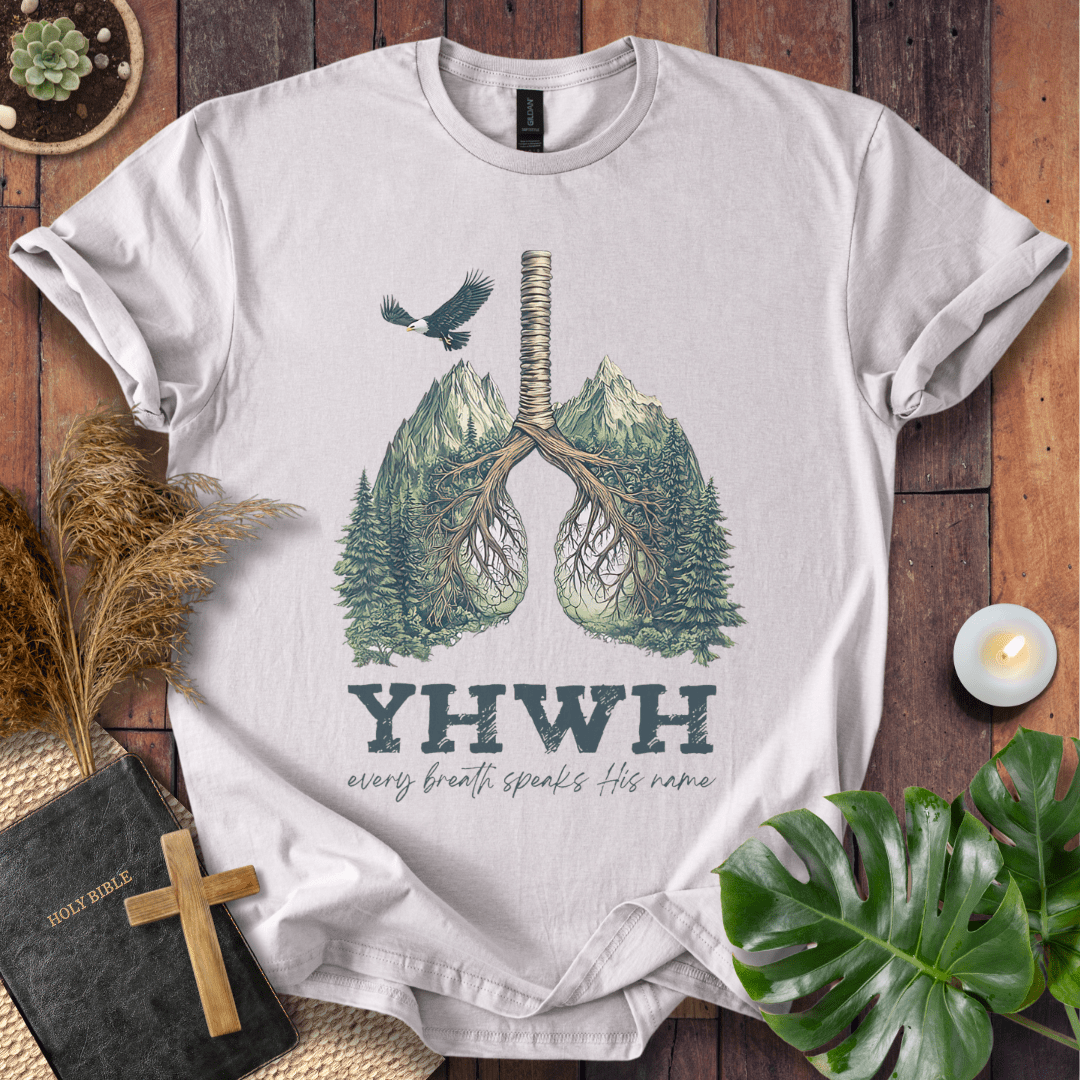 Christian t-shirt with YHWH design, tree-lung illustration, mountains, and “Every Breath Speaks His Name” text, laid flat with Bible and plants.