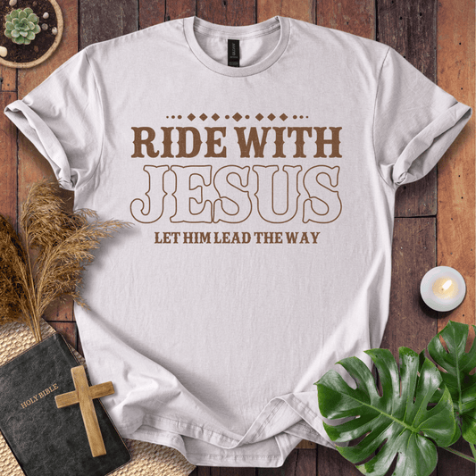 Ride With Jesus Western graphic tee on a wooden background with a Bible, cross, and green leaves.