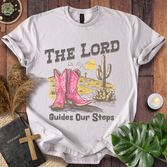 t-shirt with pink cowboy boots and "The Lord Guides Our Steps" text in a desert landscape with a cross and cactus.