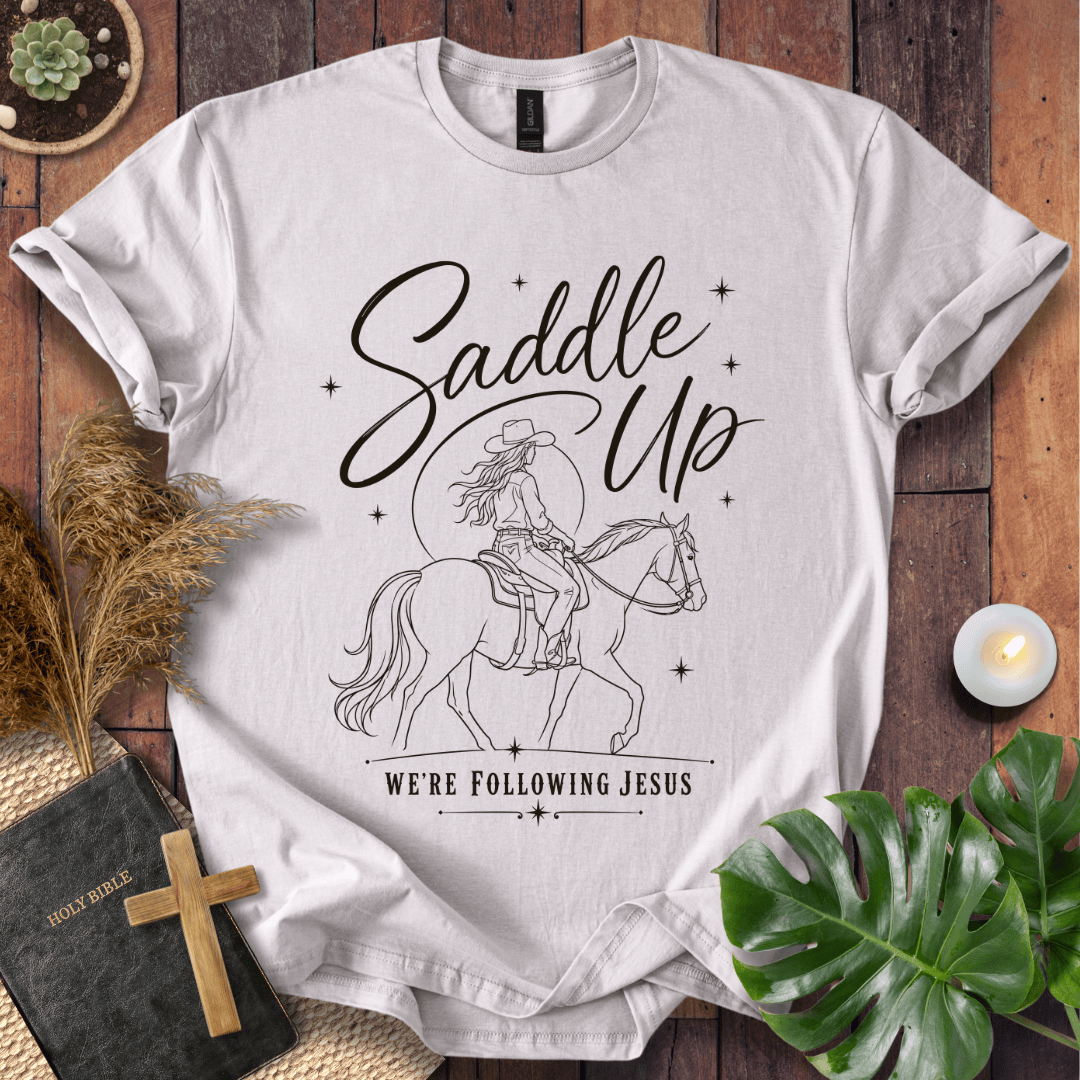 "Saddle Up, We're Following Jesus" shirt with a cowgirl riding a horse under a moonlit sky, displayed on a wooden background.