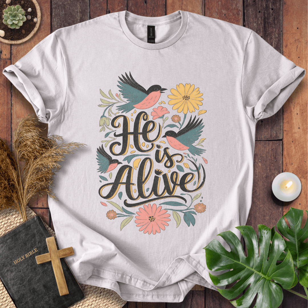 He Is Alive" Christian Easter t shirt for women with birds and flowers on a natural cream shirt, styled on a rustic wood background.