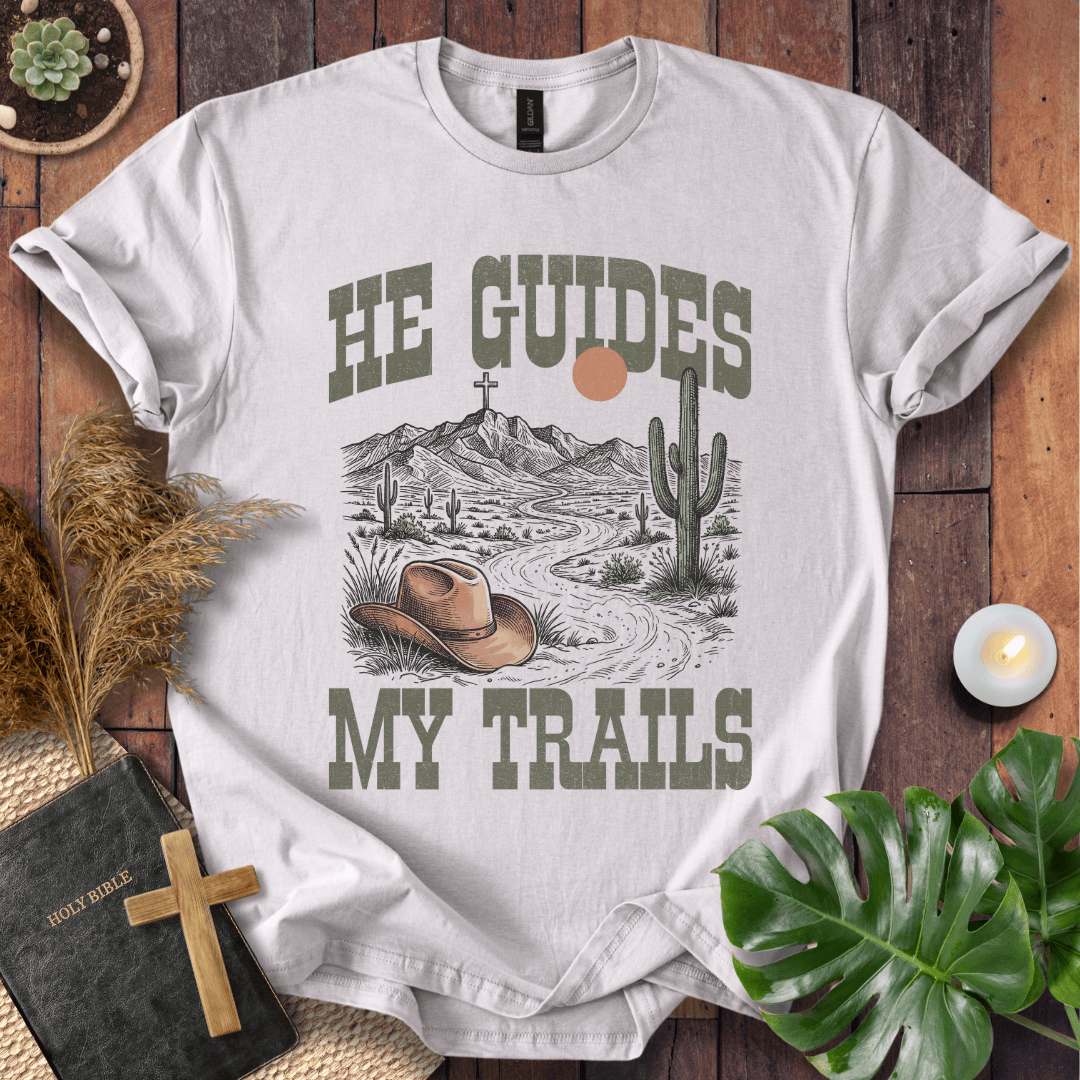 A cream-colored Christian t-shirt with a desert landscape, cowboy hat, and cross, reading He Guides My Trails.