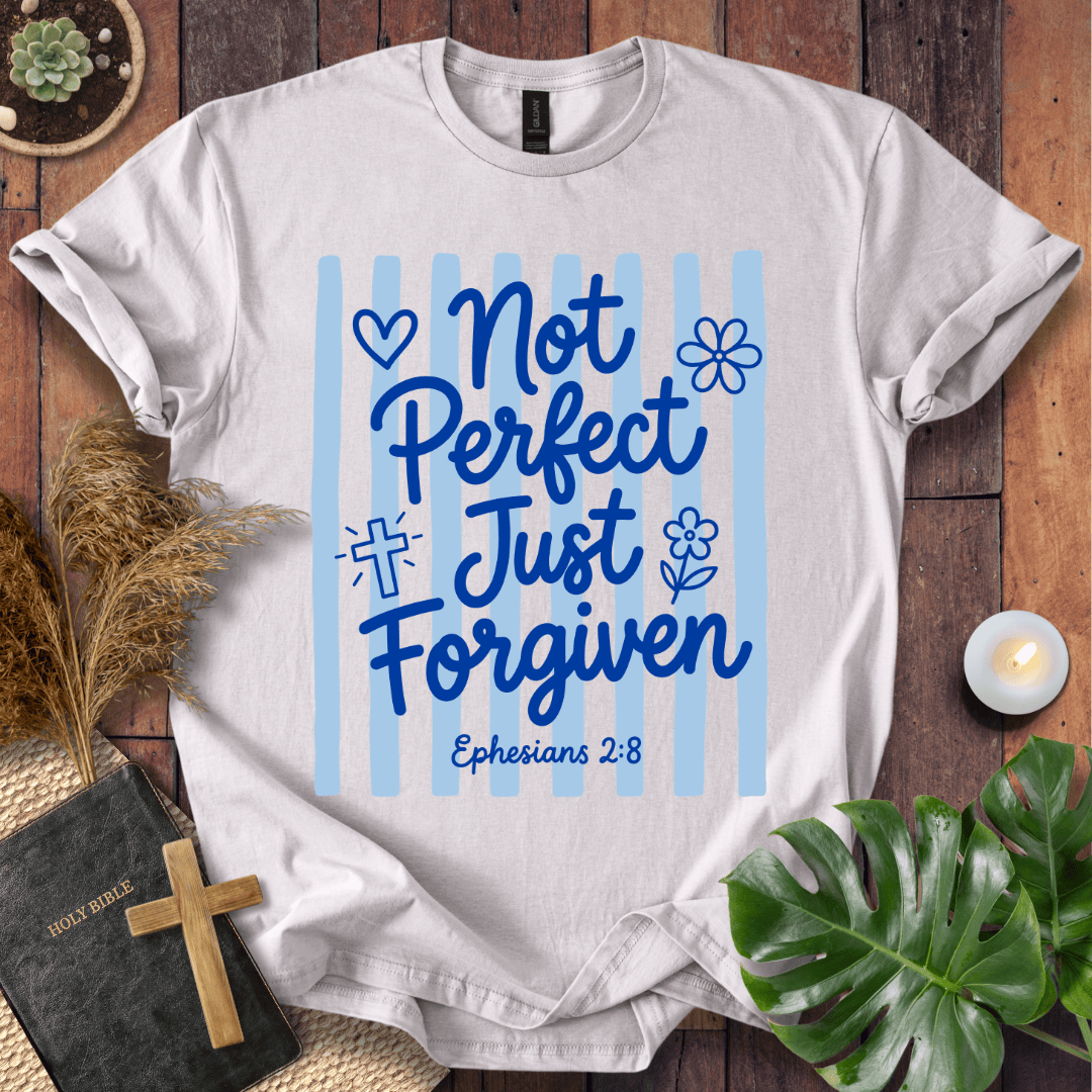 A white short-sleeve T-shirt laid flat on a wooden surface, featuring a blue graphic that reads 'Not Perfect Just Forgiven' with 'Ephesians 2:8' below, surrounded by decorative props including a book, candle, dried grass, and green leaves.