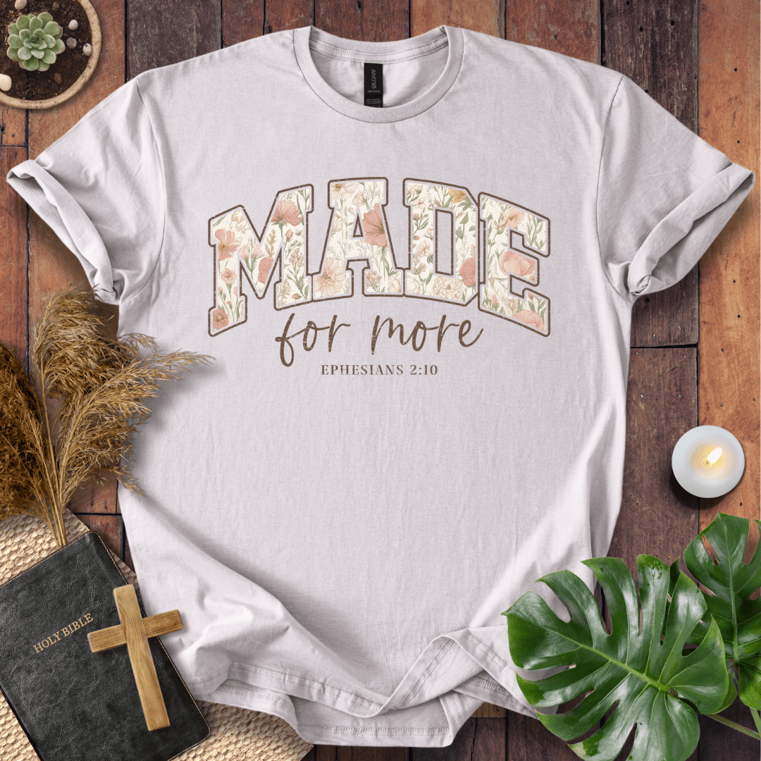 A light gray graphic t-shirt laid flat on a rustic wooden background, surrounded by decorative props including a Bible with a wooden cross, pampas grass, a small candle, and green leaves. The shirt features the text 'MADE for more EPHESIANS 2:10' in a floral patterned font.