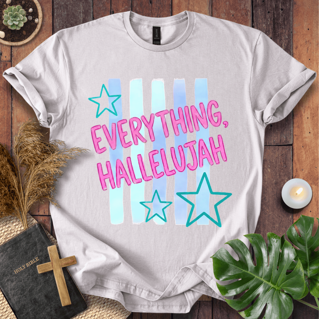A light gray graphic t-shirt laid flat on a rustic wooden surface, surrounded by decorative props including a Bible with a wooden cross, pampas grass, a small candle, and green leaves. The t-shirt features a colorful graphic with the text 'EVERYTHING, HALLELUJAH' in pink outlined letters over pastel blue and light blue vertical stripes, accented with teal stars.