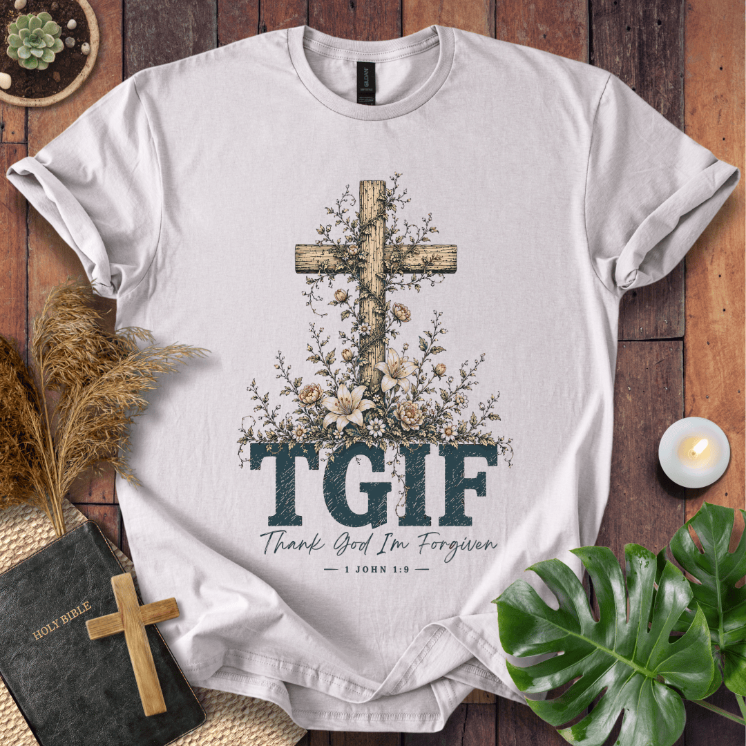 A white short-sleeve T-shirt laid flat on a rustic wooden surface, featuring a large wooden cross decorated with flowers and the text 'TGIF' with 'Thank God I'm Forgiven' and '1 John 8:32' below. The shirt is styled with a book, candle, dried grass, green leaves, and a small wooden cross.