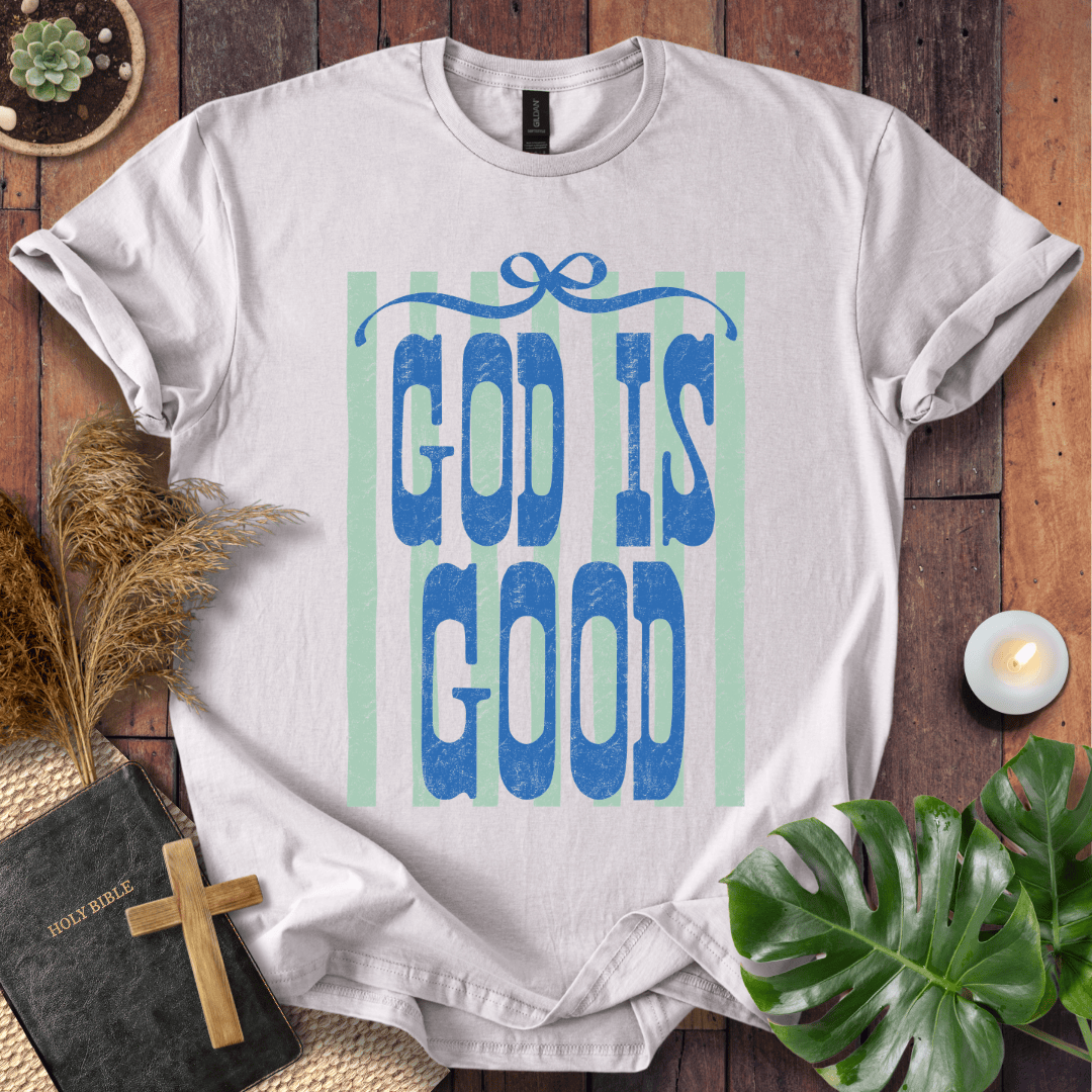 A white short-sleeve T-shirt laid flat on a rustic wooden surface, featuring the phrase 'GOD IS GOOD' in blue textured lettering with pale green vertical stripes behind it. The shirt is styled with a black Bible, a wooden cross, dried grass, a small candle, and green leaves around it.