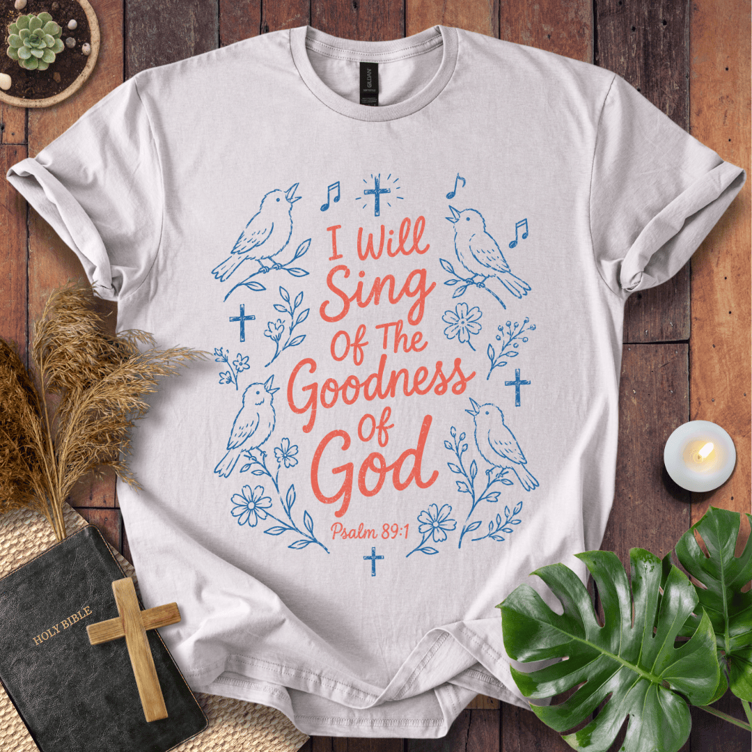 A white short-sleeve T-shirt laid flat on a rustic wood surface, decorated with blue line drawings of birds, flowers, and crosses, plus coral-red text reading 'I Will Sing Of The Goodness Of God' and 'Psalm 89:1.' Surrounding props include a Bible with a wooden cross, dried grass, a small candle, and green leaves.