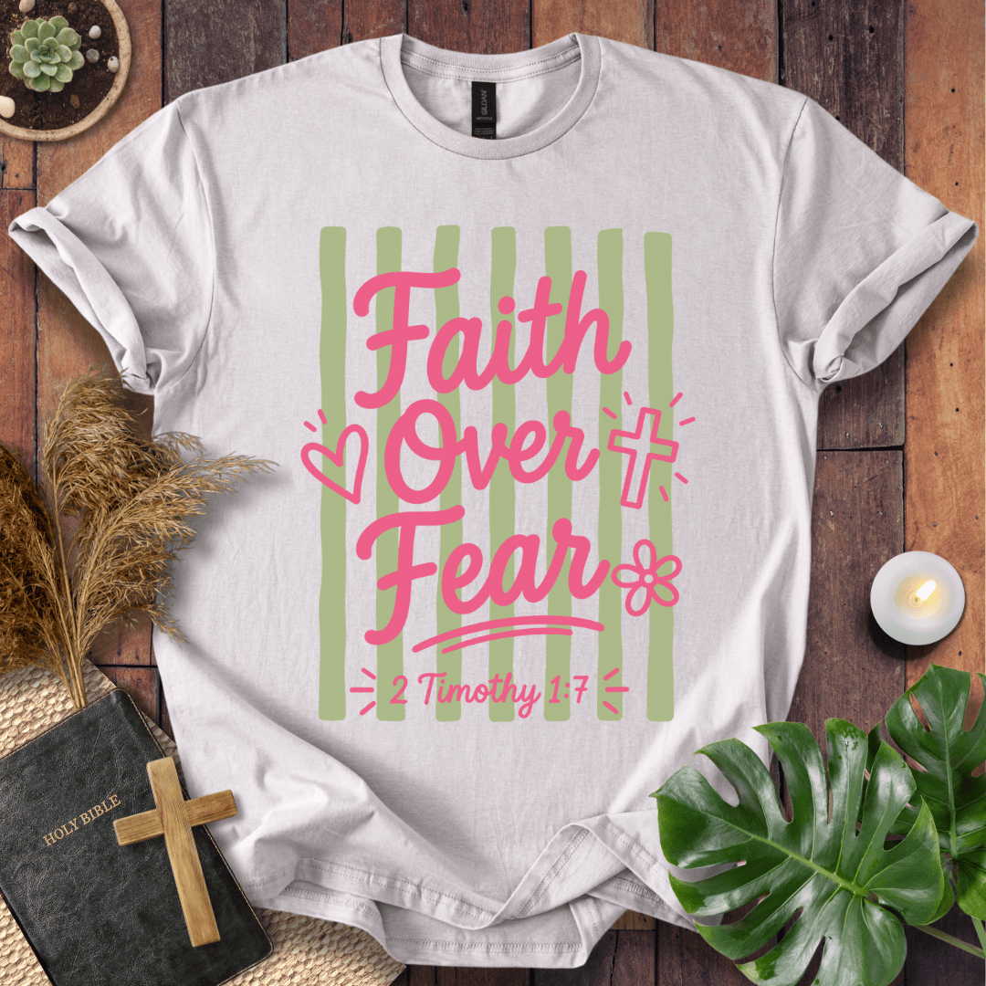 A white short-sleeve graphic T-shirt laid flat on a rustic wood surface, surrounded by decorative props including a Bible, wooden cross, dried grass, candle, and green leaves. The shirt has a large front print with vertical light green stripes, pink script text reading 'Faith Over Fear,' small pink hearts and flowers, and smaller pink text reading '2 Timothy 1:7'.