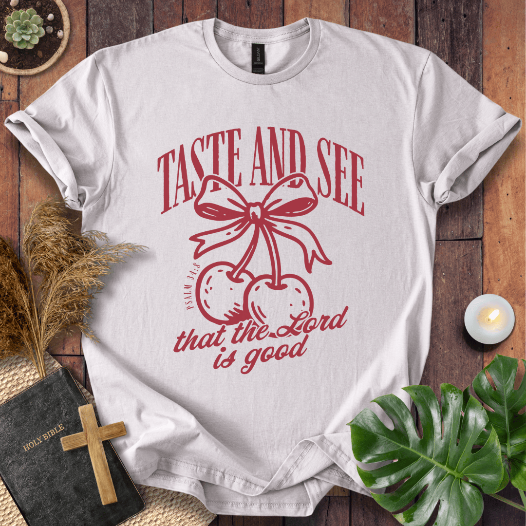 A white short-sleeve graphic T-shirt laid flat on a rustic wooden surface, featuring red text that reads 'TASTE AND SEE that the Lord is good' above a red bow and cherries design. Decorative props including a small plant, dried grass, a lit candle, a black Bible with a wooden cross, and green leaves surround the shirt.