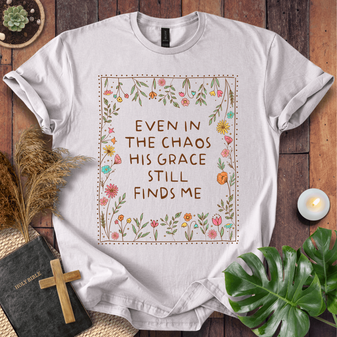 A white graphic t-shirt laid flat on a dark wood background, surrounded by decorative props including a Bible, wooden cross, dried grass, a small candle, and green leaves. The t-shirt features a central floral border with the text 'EVEN IN THE CHAOS HIS GRACE STILL FINDS ME'.