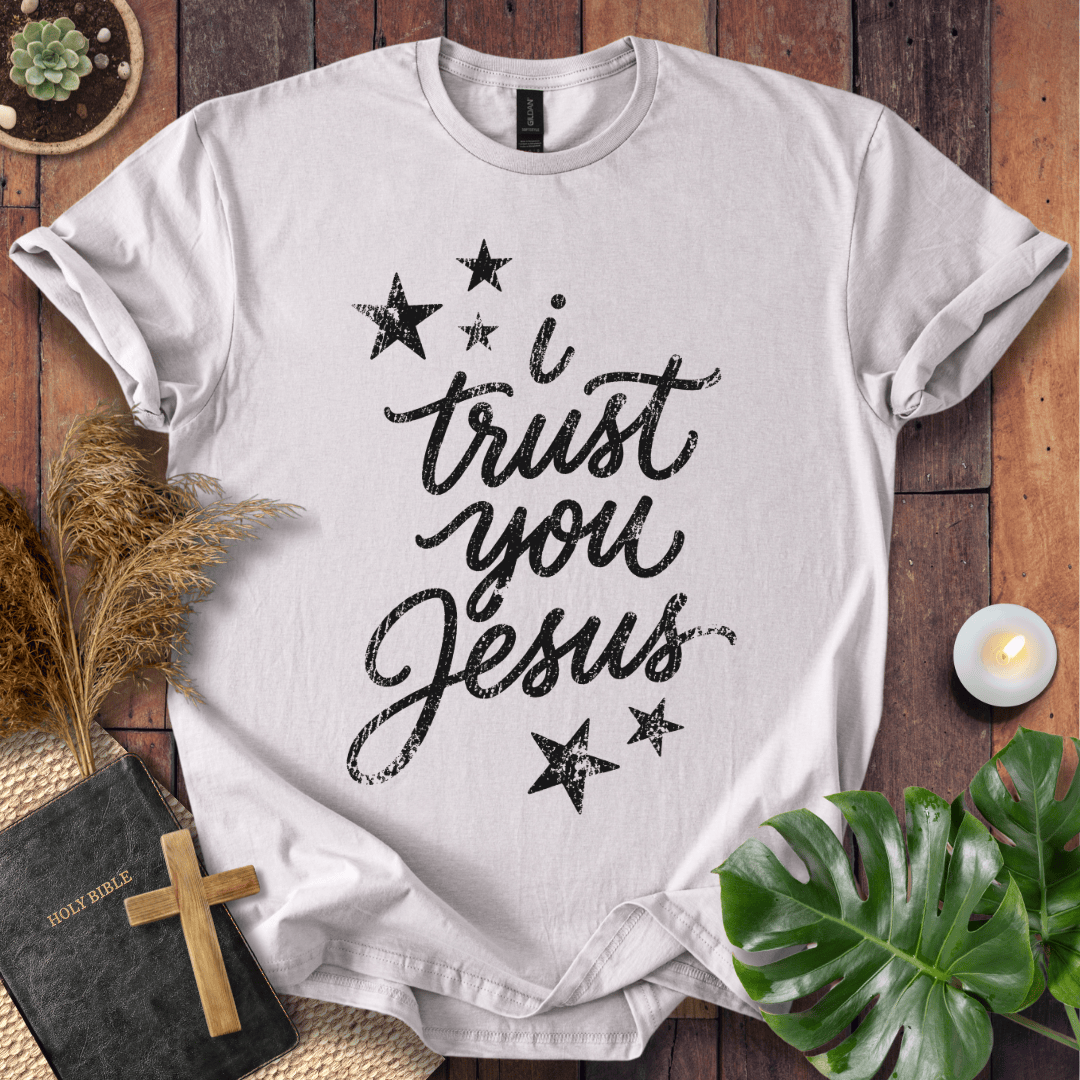 A white short-sleeve T-shirt laid flat on a rustic wooden surface, printed with the phrase 'I trust you Jesus' in black script and surrounded by black stars. The shirt is styled with a Bible, wooden cross, dried grass, green leaves, and a small candle.