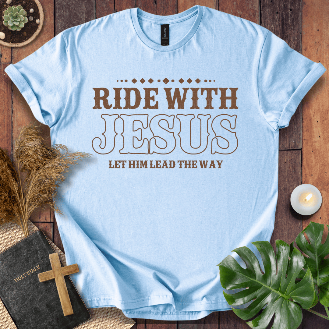 Ride With Jesus Western graphic tee on a wooden background with a Bible, cross, and green leaves.