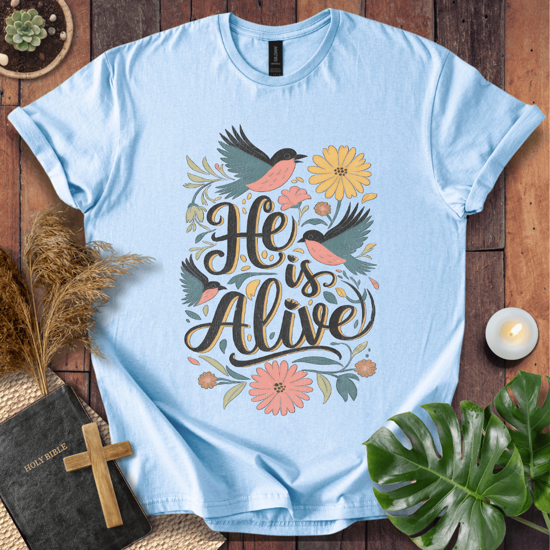 He Is Alive" Christian Easter t shirt for women with birds and flowers on a natural cream shirt, styled on a rustic wood background.