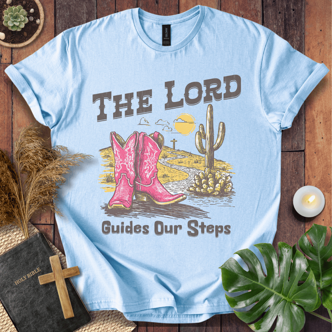t-shirt with pink cowboy boots and "The Lord Guides Our Steps" text in a desert landscape with a cross and cactus.