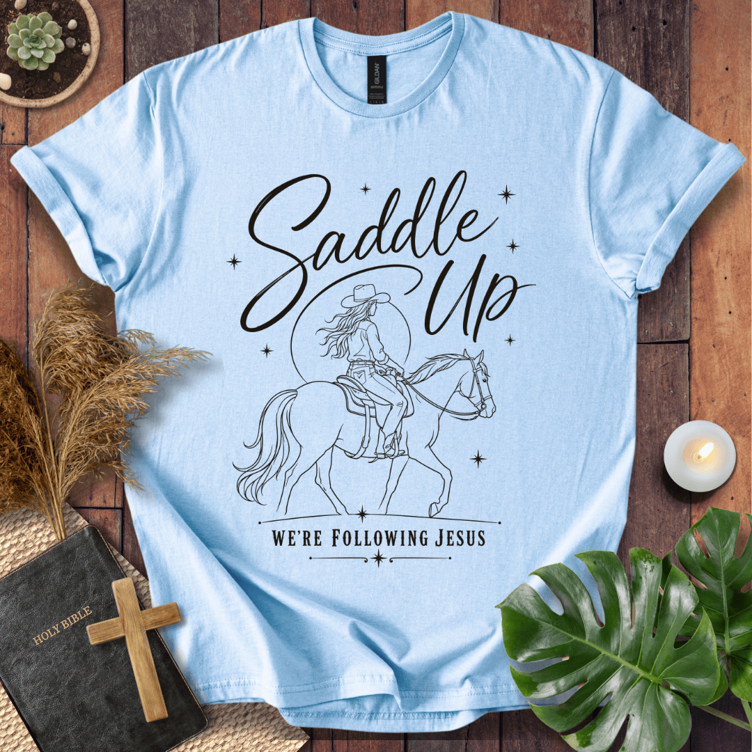 "Saddle Up, We're Following Jesus" shirt with a cowgirl riding a horse under a moonlit sky, displayed on a wooden background.