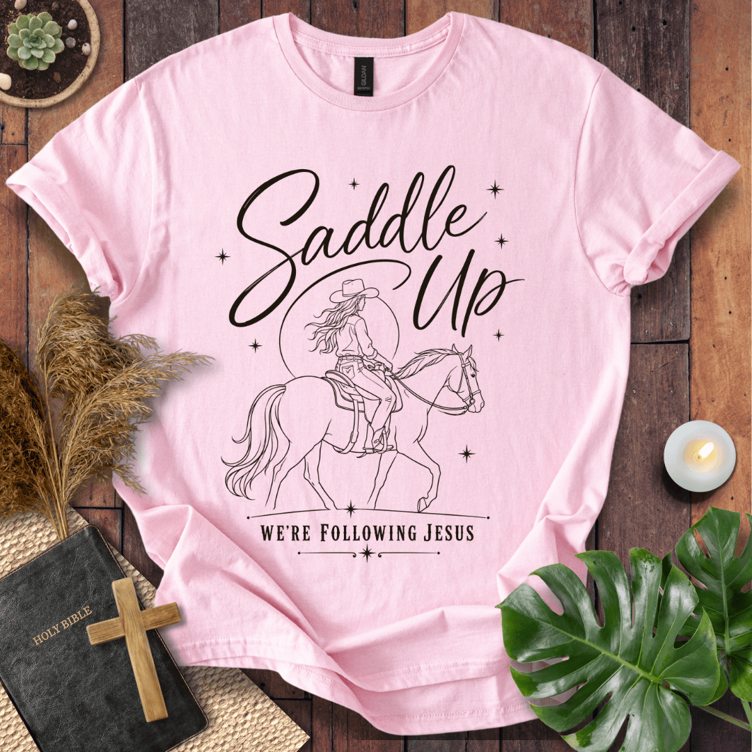 "Saddle Up, We're Following Jesus" shirt with a cowgirl riding a horse under a moonlit sky, displayed on a wooden background.