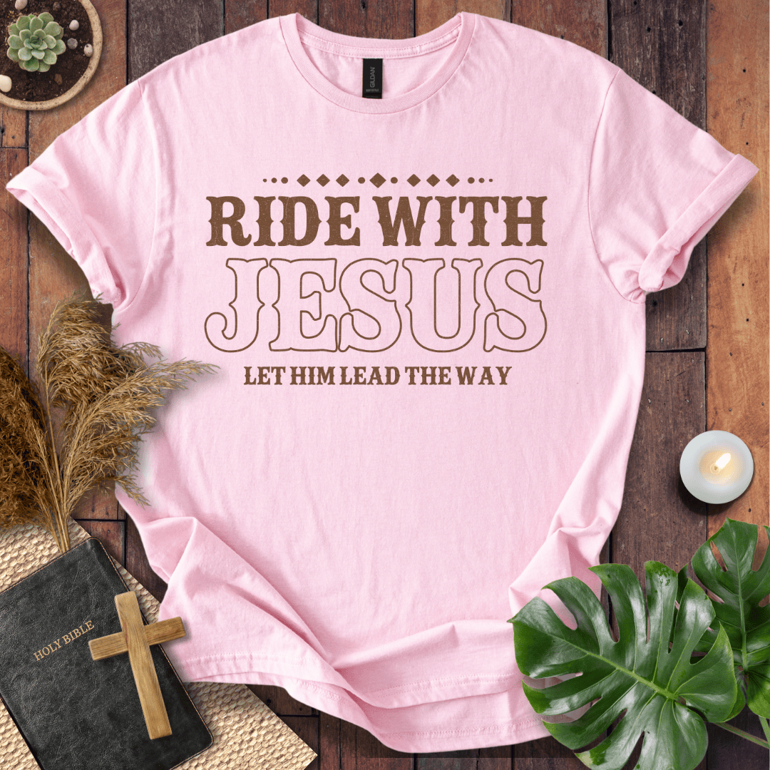 Ride With Jesus Western graphic tee on a wooden background with a Bible, cross, and green leaves.