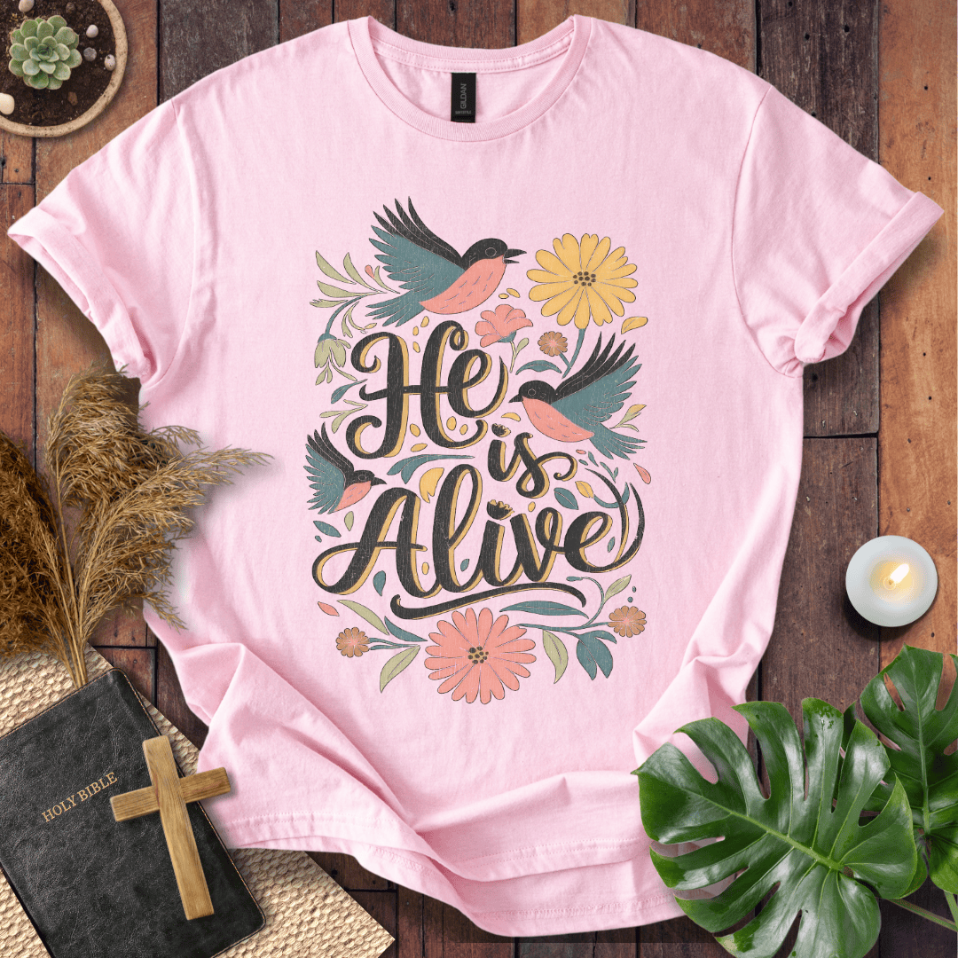 He Is Alive" Christian Easter t shirt for women with birds and flowers on a natural cream shirt, styled on a rustic wood background.