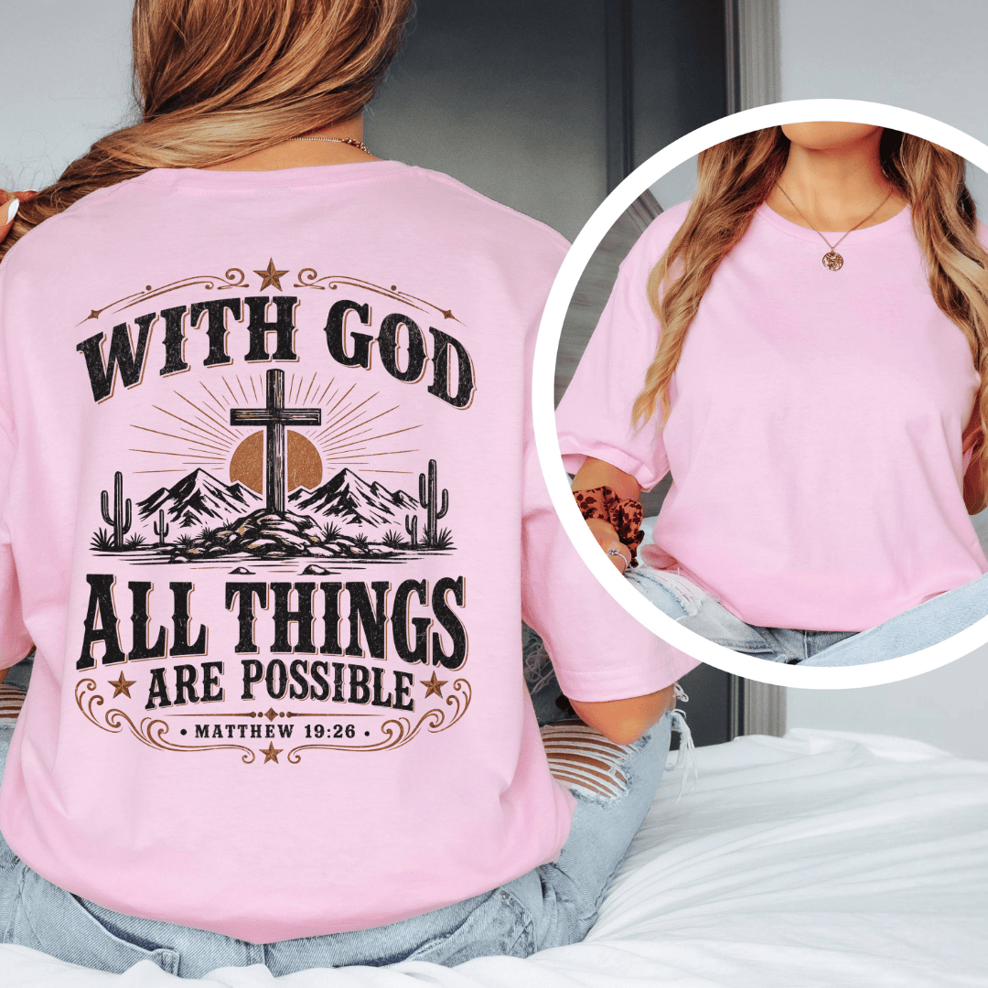 A woman sitting on a bed wearing a western faith shirt featuring a desert cross design and Matthew 19:26 scripture.