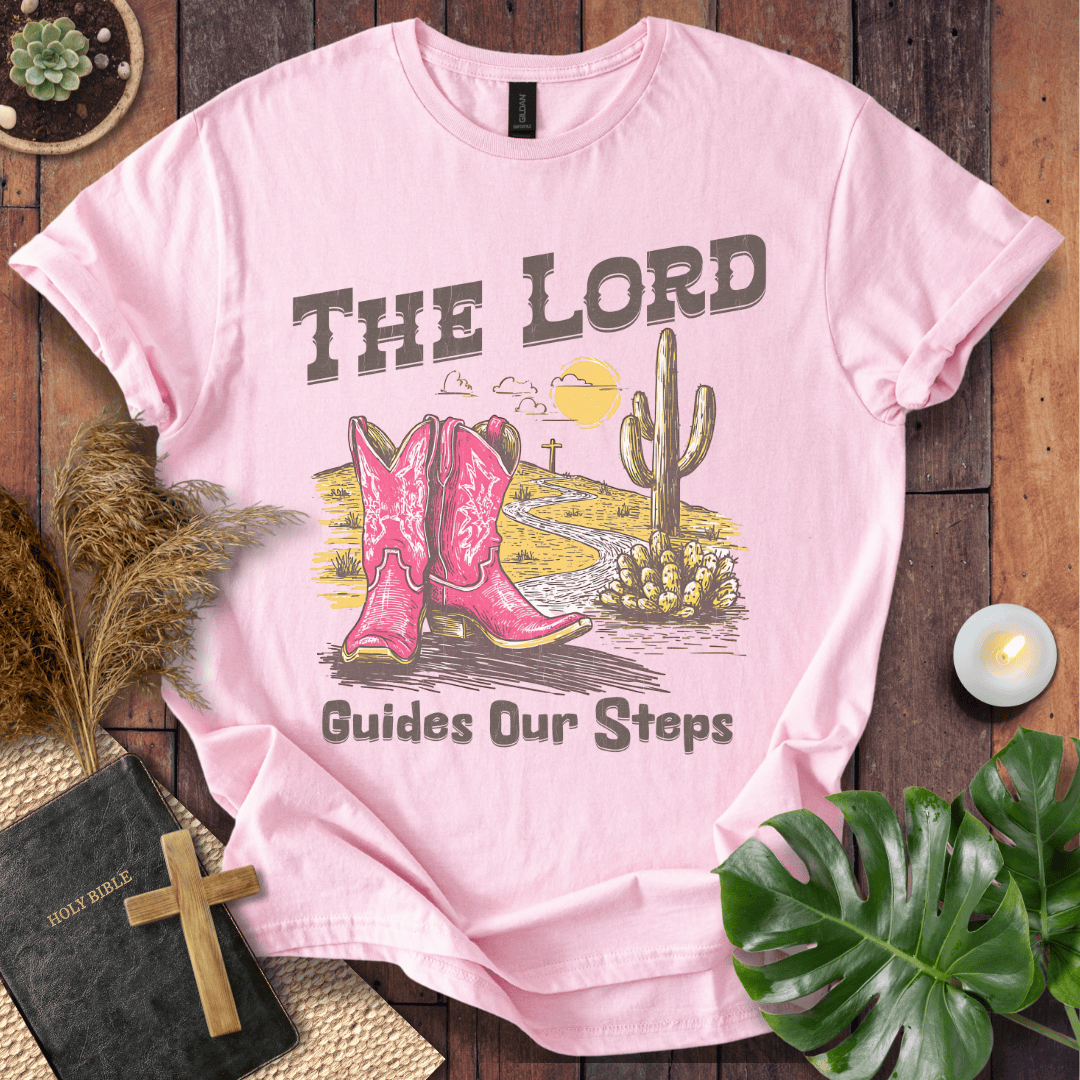 t-shirt with pink cowboy boots and "The Lord Guides Our Steps" text in a desert landscape with a cross and cactus.