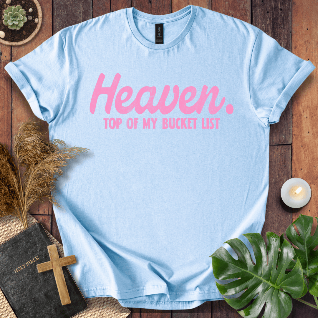 A light blue crewneck t-shirt with pink text reading 'Heaven. TOP OF MY BUCKET LIST' is displayed in a flat-lay arrangement on a wooden surface with a bible, cross, pampas grass, a monstera leaf, and a small candle.