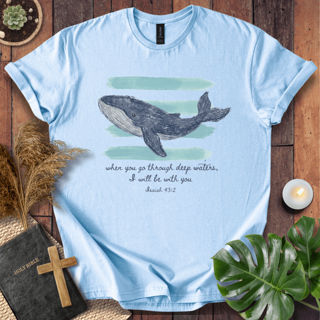 A light blue graphic T-shirt laid flat on a rustic wooden surface, featuring a watercolor-style whale illustration above the text, 'when you go through deep waters, I will be with you Isaiah 43:2.' The shirt is styled with a Bible, wooden cross, dried grass, green leaves, and a small candle.