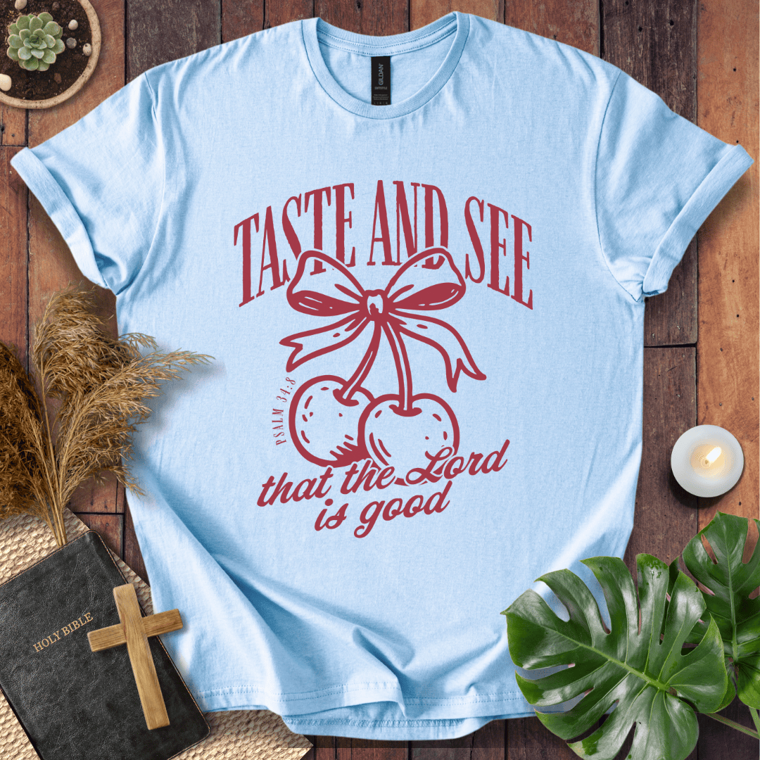A light blue crewneck t-shirt with a red graphic of cherries under a bow and the text 'TASTE AND SEE that the Lord is good'. The shirt is displayed in a flat-lay arrangement on a wooden surface with a bible, cross, pampas grass, a monstera leaf, and a small candle.