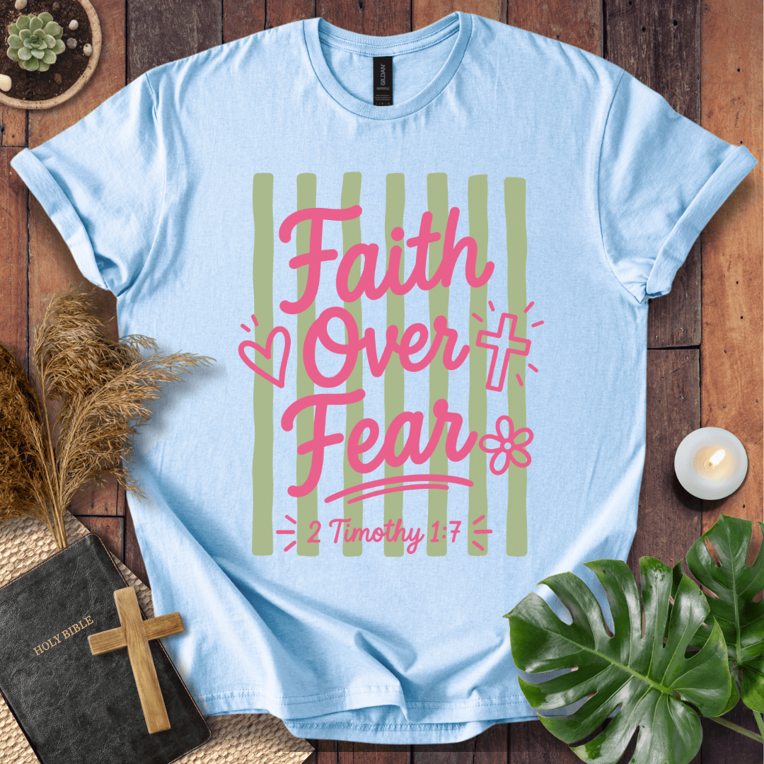 A light blue short-sleeve t-shirt with a graphic reading 'Faith Over Fear' in pink script over vertical sage green stripes, with '2 Timothy 1:7' below. The shirt is displayed in a styled flat-lay on a wooden surface with a Bible, cross, pampas grass, candle, and green leaves.