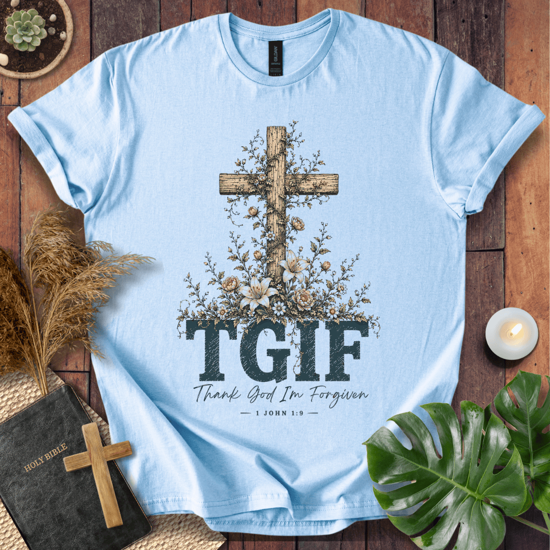 A light blue short-sleeve T-shirt laid flat on a rustic wooden surface, featuring a large wooden cross decorated with flowers and the text 'TGIF' with 'Thank God I'm Forgiven' underneath. The shirt is styled with a Bible, a small cross, dried grass, a candle, and green leaves.