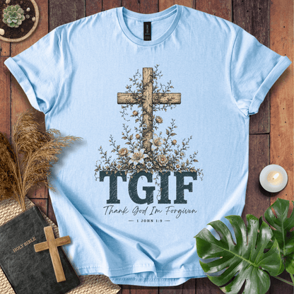 A light blue short-sleeve T-shirt laid flat on a rustic wooden surface, featuring a large wooden cross decorated with flowers and the text 'TGIF' with 'Thank God I'm Forgiven' underneath. The shirt is styled with a Bible, a small cross, dried grass, a candle, and green leaves.
