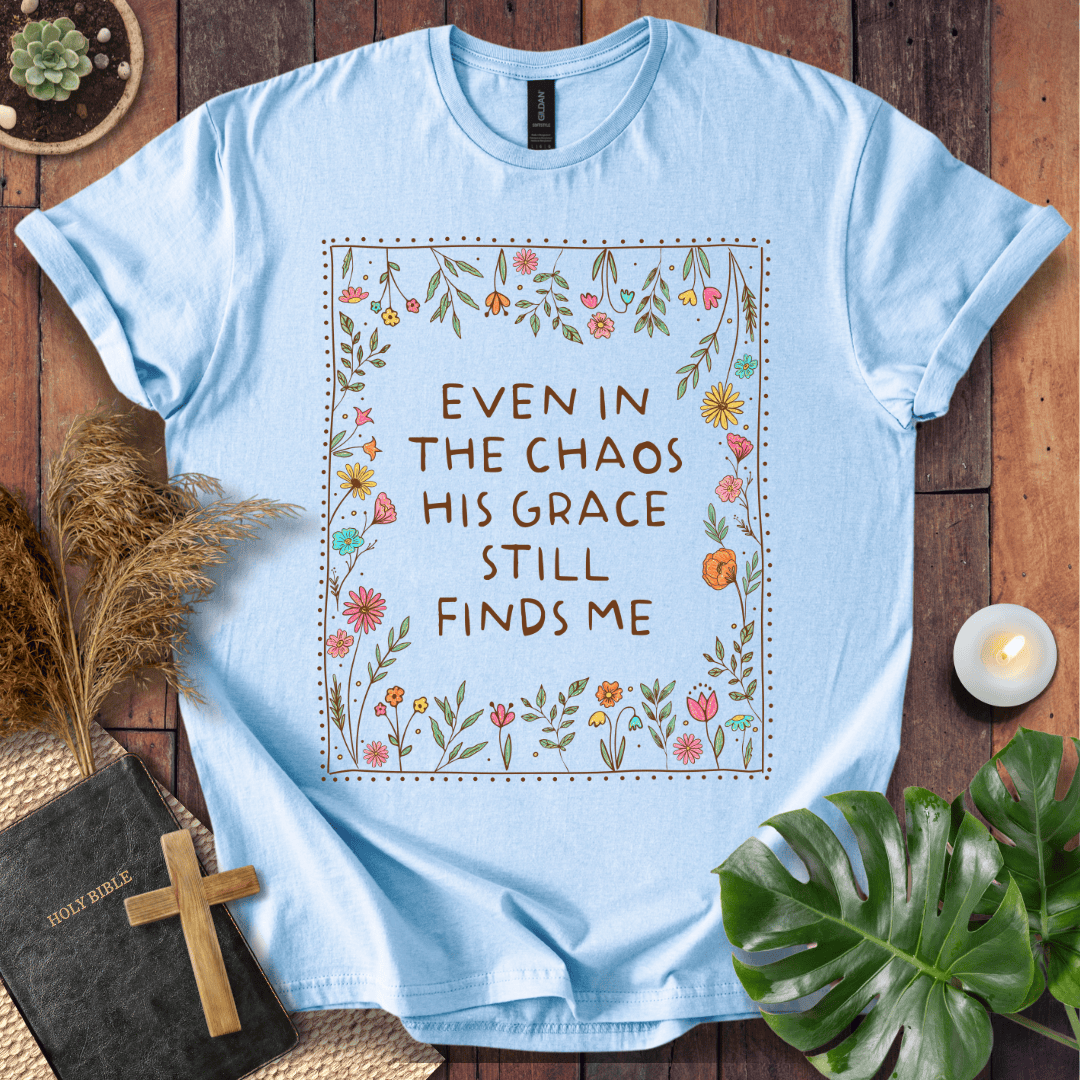 A light blue short-sleeve T-shirt laid flat on a rustic wood surface, featuring a floral border and the phrase 'EVEN IN THE CHAOS HIS GRACE STILL FINDS ME' printed on the front. Decorative props including a book with a cross, dried grass, a small candle, and green leaves surround the shirt.