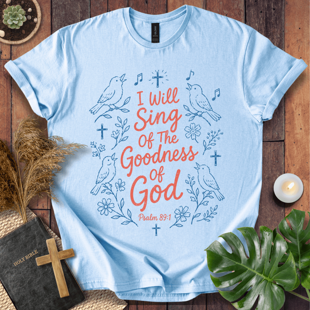 A light blue graphic T-shirt laid flat on a wooden surface, surrounded by decorative props including a Bible with a wooden cross, dried grass, a small plant, a candle, and green leaves. The shirt has a blue line-art design with birds, flowers, music notes, and the text 'I Will Sing Of The Goodness Of God' and 'Psalm 85:1' in coral and blue lettering.